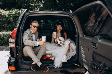 Gelin ve damat bir arabanın bagajında oturmuş biraz yemek yiyor, kola içiyor ve hamburger yiyor. 