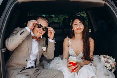 Gelin, şapka ve damat takıp bir arabanın bagajında oturmuş patates kızartması yerken eğleniyor. 