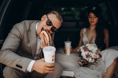 Gelin ve damat bir arabanın bagajında oturmuş biraz yemek yiyor, kola içiyor ve hamburger yiyor. 