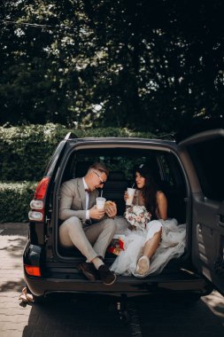 Gelin ve damat bir arabanın bagajında oturmuş biraz yemek yiyor, kola içiyor ve hamburger yiyor. 