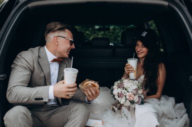 Gelin ve damat bir arabanın bagajında oturmuş biraz yemek yiyor, kola içiyor ve hamburger yiyor. 