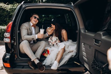 Gelin ve damat bir arabanın bagajında oturup biraz yemek yer, kola içer ve hamburgerlerini paylaşır. 