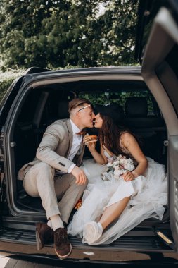 Gelin şapkalı ve damadı arabanın bagajında oturmuş hamburger yiyor ve öpüşüyor.  
