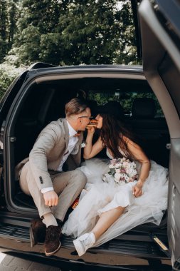 Gelin, kep ve damat takmış bir arabanın bagajında oturmuş hamburgerini paylaşıyor. 