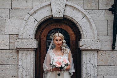 Bir buket çiçekli güzel sarışın gelin eski binanın yanında duruyor.