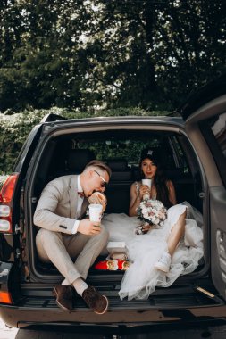 Gelin ve damat bir arabanın bagajında oturmuş biraz yemek yiyor, kola içiyor ve hamburger yiyor. 