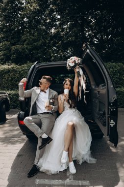 Güzel bir çift yeni evli arabada poz veriyor kola içiyor, damat geline pazılarını gösterirken eğleniyor, buketini kaldırıyor.