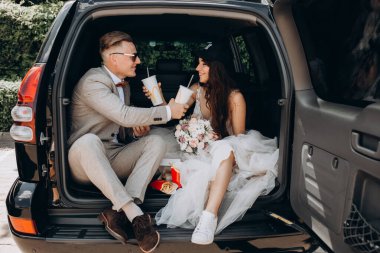 Gelin ve damat bir arabanın bagajında oturmuş biraz yemek yiyor, kola içiyor.