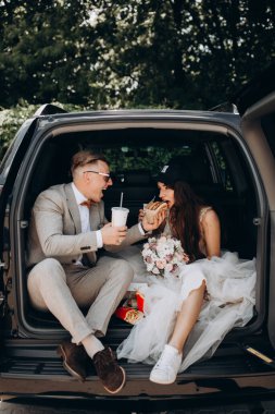 Gelin ve damat bir arabanın bagajında oturmuş biraz yemek yiyor, kola içiyor ve hamburger yiyor. 