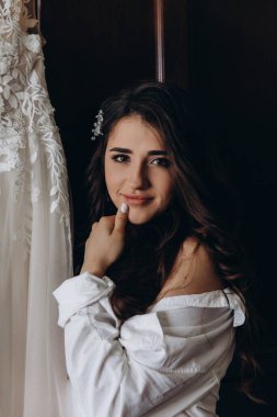 Beyaz gömlekli güzel gelinin portresi gelinliğin yanında poz veriyor. düğün sabahı