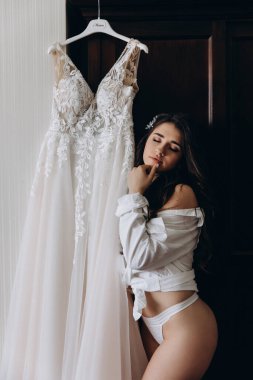 Gecelik ve beyaz gömlekli güzel gelinin portresi gelinliğin yanında poz veriyor. düğün sabahı
