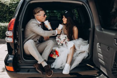 Gelin ve damat bir arabanın bagajında oturmuş biraz yemek yiyor, kola içiyor.