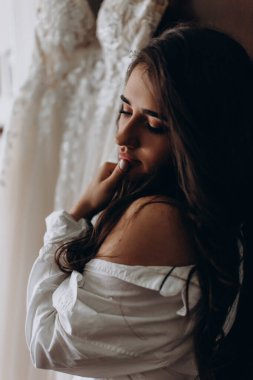 Beyaz gömlekli güzel gelinin portresi gelinliğin yanında poz veriyor. düğün sabahı