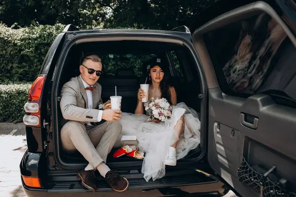 Gelin ve damat bir arabanın bagajında oturmuş biraz yemek yiyor, kola içiyor ve hamburger yiyor. 