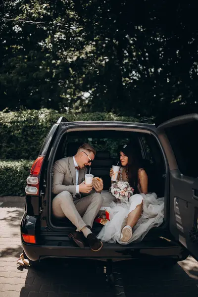 Gelin ve damat bir arabanın bagajında oturmuş biraz yemek yiyor, kola içiyor ve hamburger yiyor. 