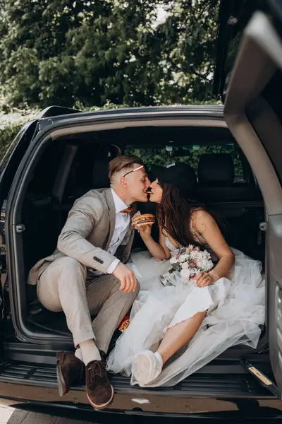 Gelin şapkalı ve damadı arabanın bagajında oturmuş hamburger yiyor ve öpüşüyor.  