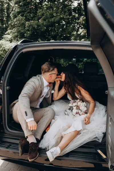 Gelin, kep ve damat takmış bir arabanın bagajında oturmuş hamburgerini paylaşıyor. 