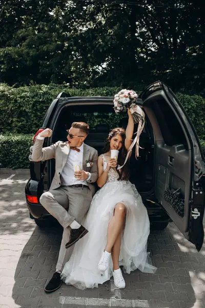 Güzel bir çift yeni evli arabada poz veriyor kola içiyor, damat geline pazılarını gösterirken eğleniyor, buketini kaldırıyor.