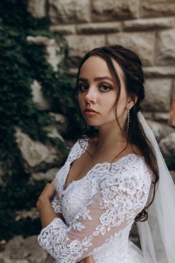 Beyaz gelinlik ve duvak giymiş, kollarını kavuşturup kameraya bakan gelin.