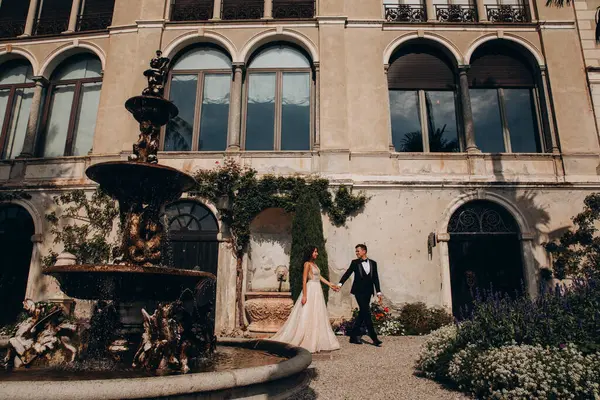 Güzel, mutlu bir düğün çifti el ele tutuşuyor ve eski evin etrafında yürüyor.  
