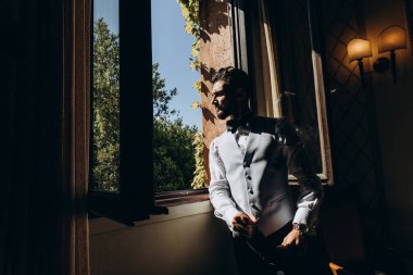 Takım elbiseli yakışıklı damat pencere eşiğine eğiliyor ve pencereye bakıyor.