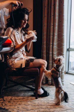 Genç bir kadın sandviç yiyor ve kuaför saç stili yaparken küçük sevimli bir köpeğe bakıyor.