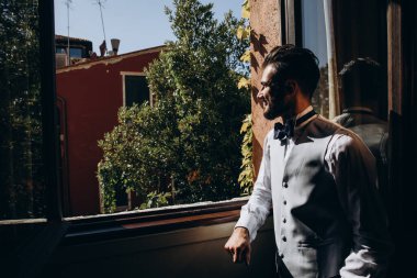 Zarif kıyafetli genç adam pencere eşiğine eğiliyor ve sabahları pencereye bakıyor.       