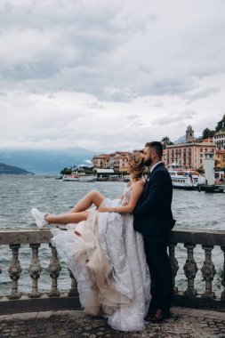 Gelin ve damat eski kasabanın arka planında göl kenarında kucaklaşıyorlar.