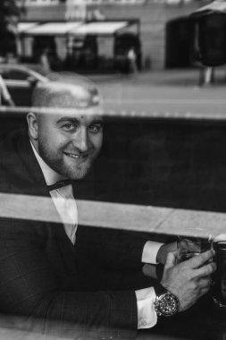 Lokantada gülümseyen ve cam tutan bir adamın siyah beyaz görüntüsü. Pencereden dışarı bakıyor. 