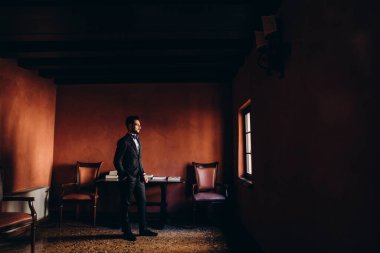 Siyah takım elbiseli bir adam odada dikiliyor ve başka tarafa bakıyor.