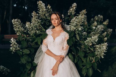 Parktaki yeşil bitkilerin arka planında poz veren beyaz elbiseli güzel gelin.