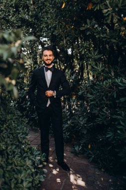 Takım elbiseli yakışıklı adam parkta duruyor ve düğün günü kameraya gülümsüyor.
