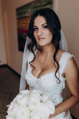 Elinde buketi olan güzel bir gelin. İçeride dikilip kameraya bakıyor.