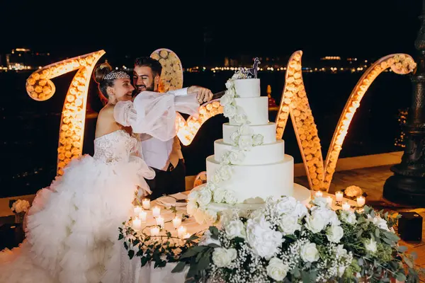 Düğün çifti gülüyor ve düğün pastasını kesiyor.  