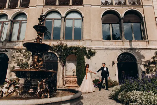 Güzel, mutlu bir düğün çifti el ele tutuşuyor ve eski evin etrafında yürüyor.  