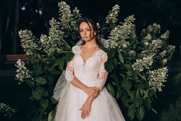 Parktaki yeşil bitkilerin arka planında poz veren beyaz elbiseli güzel gelin.