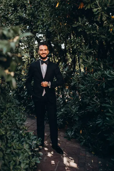Takım elbiseli yakışıklı adam parkta duruyor ve düğün günü kameraya gülümsüyor.