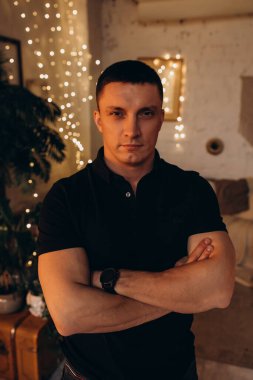 Ciddi ifadeli çekici bir adam, kollarını kavuşturmuş, Noel 'i Noel süslemesiyle dolu bir odada geçiriyor. 