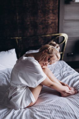 Yatak odasında beyaz ipek bornozlu çekici sarışın kadın.                