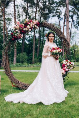 Güzel genç gelinin portresi düğün kemerinin yanında bir buket çiçek tutuyor.     