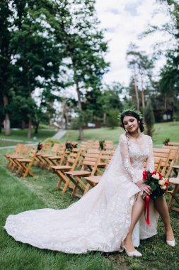 Güzel genç gelinin portresi düğün günü dışarıda, tahta sandalyede oturuyor.       