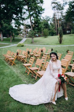 Güzel genç gelinin portresi düğün günü dışarıda, tahta sandalyede oturuyor.       