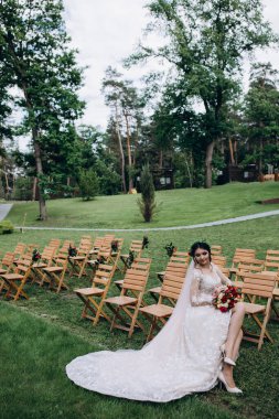 Güzel genç gelinin portresi düğün günü dışarıda, tahta sandalyede oturuyor.       