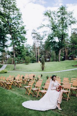 Güzel genç gelinin portresi düğün günü dışarıda, tahta sandalyede oturuyor.       
