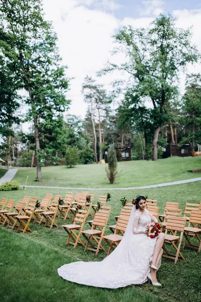 Güzel genç gelinin portresi düğün günü dışarıda, tahta sandalyede oturuyor.       