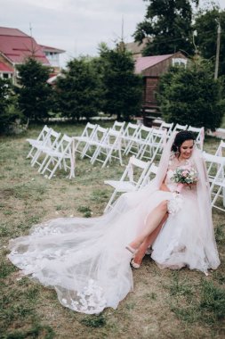 Gelinlik giymiş güzel bir gelin, elinde çiçekler, sandalyede oturuyor.