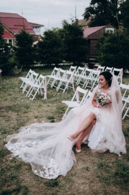 Gelinlik giymiş güzel bir gelin, elinde çiçekler, sandalyede oturuyor.