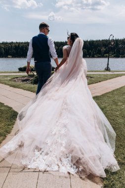 Mutlu genç gelin ve damadın el ele tutuşup göl kenarında yürüdüğü arka manzara.