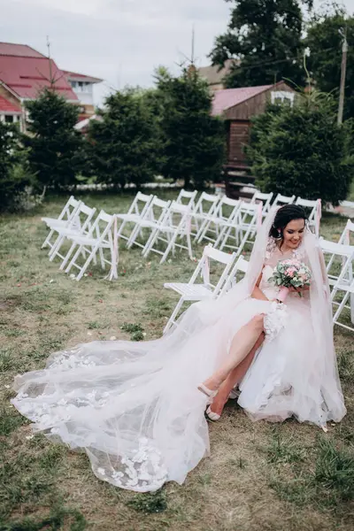 Gelinlik giymiş güzel bir gelin, elinde çiçekler, sandalyede oturuyor.