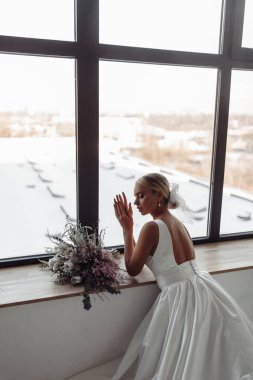 Pencere eşiğine güzel buketlerle yaslanmış gelinlik giymiş çekici genç bir gelin.          